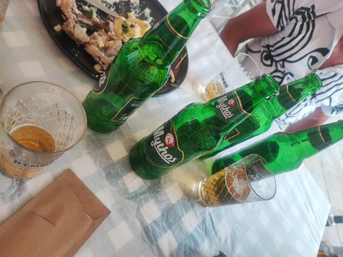 Tre ölflaskor och två halvfulla glas på ett rutigt bordstyg, rester av en måltid i bakgrunden.