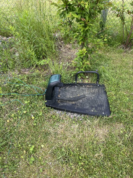 PARKSIDE-maskin på gräs intill laddplatta, felkopplad och ej fungerande, med trädgård i bakgrunden.