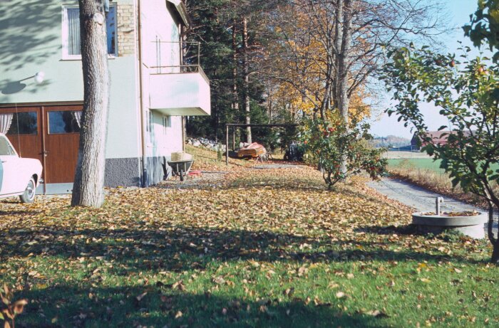 Ett vintage foto av ett hus från 1965 med en gammal bil parkerad, omgivet av lövträd och en gräsmatta täckt av löv.