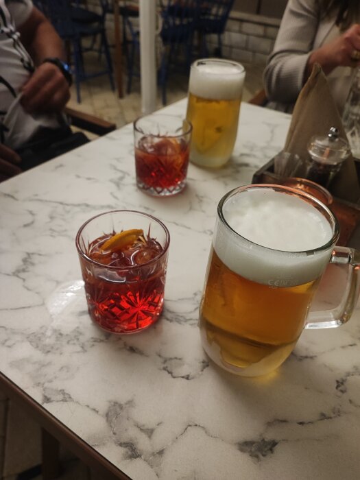 Drycker på marmorbord: en öl, två glas med rött innehåll, personer i bakgrunden.