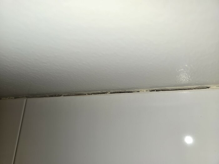 Delvis borttagen silikonfog mellan vitt kakel och målat tak i ett badrum, rester av silikon syns kvar.
