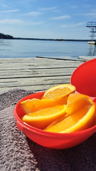 Apelsinskivor i röd skål på en brygga vid vatten med soligt väder.