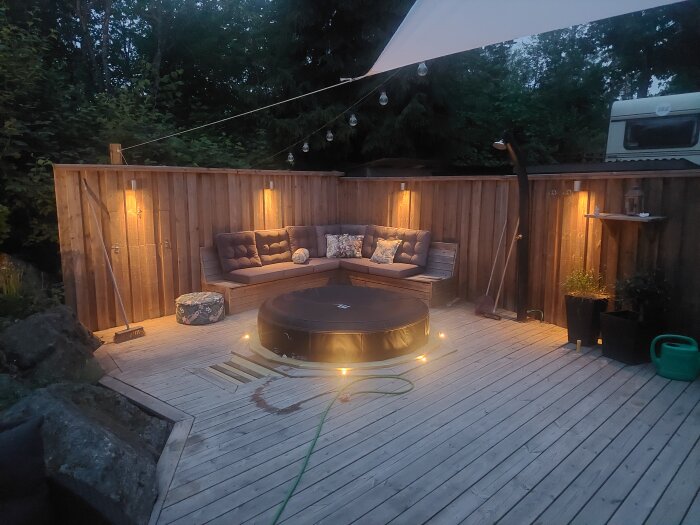 Rund, uppblåsbar spa-pool på ett trädeck med soffhörna och ljusslinga i en trädgård vid skymning.