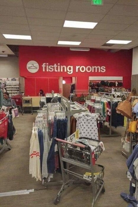 Interiör av klädaffär med klädställningar och ett felstavat skylt för 'fitting rooms' som ser ut att stå 'fisting rooms'.