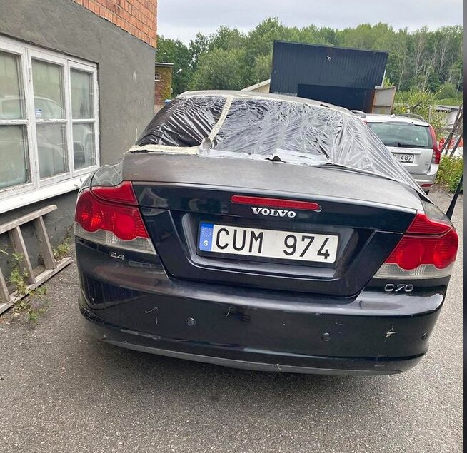 Svart Volvo C70 med trasig bakruta täckt med plastfolie.