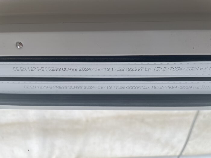Stängningskod tryckt på kanten av ett glasfönster, med datum och tillverkarinformation.