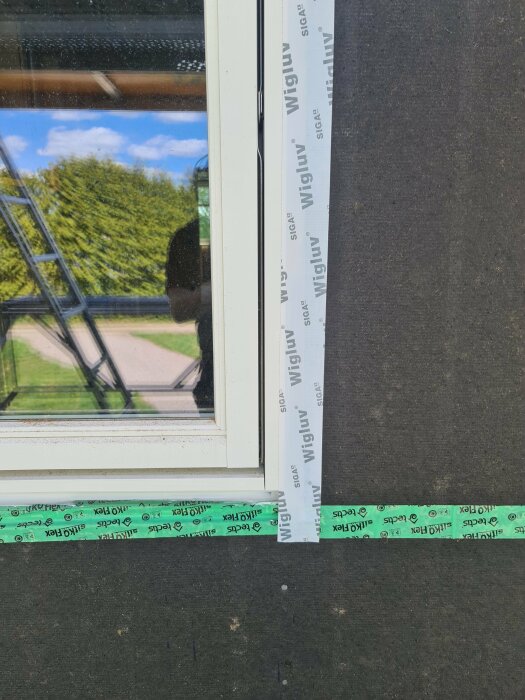 Tejpning mellan fönsterkarm och regel med wigluv-tape samt en spegling av trädgården i fönstret.
