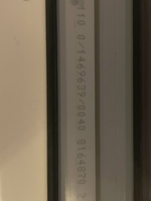 Närbild på en etikett med serienummer på en byggdels kant, foto med dålig belysning.