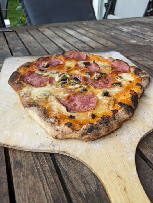 Hemlagad pizza med salami och champinjoner på träbräda ovanpå ett träbord.