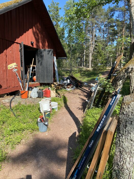 Röd stuga med öppna dörrar och byggmaterial utspritt runtomkring på en solig dag.