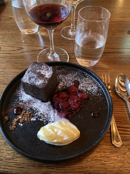 Efterrätt med chokladkaka, bär och kräm på svart tallrik, flankerad av vin och vattenglas.