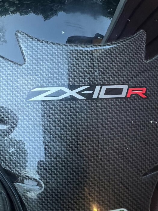 Logotypen "ZX10R" i vit och röd text på en svart kolfiberbakgrund på en motorcykeldel.