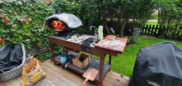 Hemmagjort bord av cortenstål med skifferplattor, bakugn och matlagningsredskap i en trädgård.