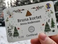 Hand håller 'Bruna kortet' certifiering med namn och illustrationer på natur och bajs.