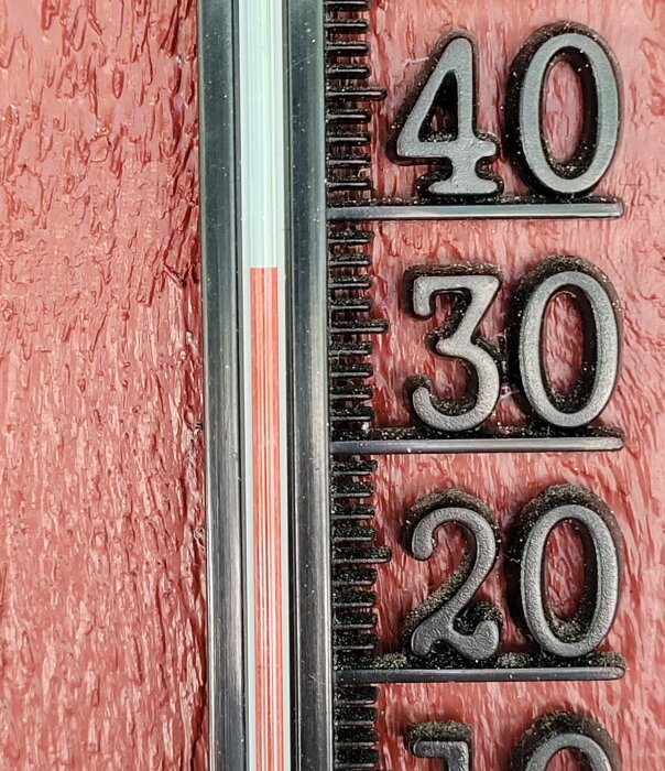 Termometer visar nästan 35 grader Celsius mot röd trävägg.