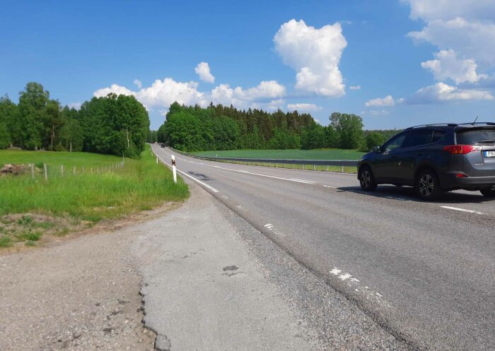 Landsväg nära betesmark med förbipasserande bil och tydlig vägmärkning, omgivet av gröna fält och skog.