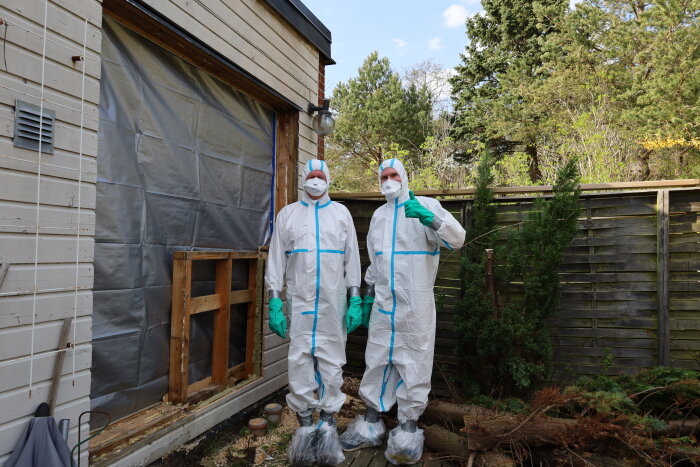 Två personer i skyddsoveraller framför hem med plasttäckt öppning där altandörrar tagits bort.