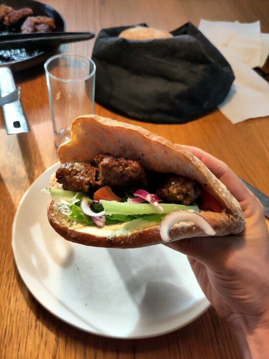 Hand som håller cevapi i bröd med sallad och lök, serverat på en vit tallrik.