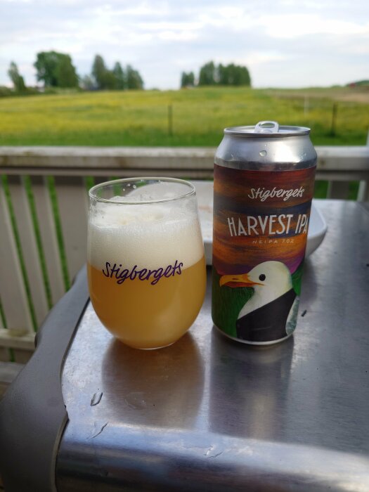 Ölglas och burk med etiketten "Harvest IPA" på ett bord utomhus, landskap i bakgrunden.