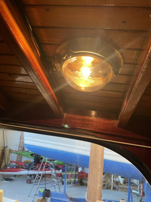 Mässingstakarmatur monterad i mahognybåt med tänd lampa, synlig träruff och utsikt till verkstad genom fönstret.