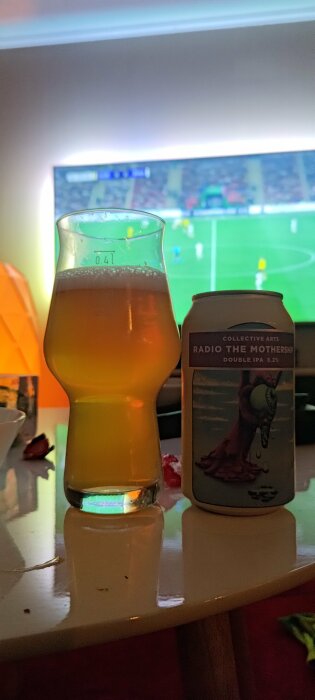 Ölglas och burk av Radio The Mothership IPA framför en fotbollsmatch på TV.