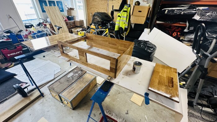 Snickeriprojekt med en halvfärdig träram för ett förarsäte och snickerimaterial i en verkstad med verktyg och cyklar i bakgrunden.
