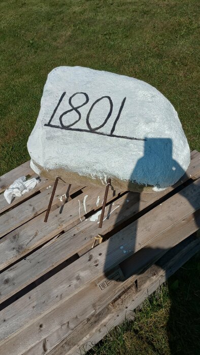 Målad sten med husnummer 1801 på träpall och målarföremål runtomkring.