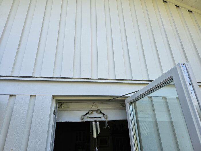 Vitmålad lockpanel på en husvägg med en öppen dörr och en ovanliggande plåtanslutning som söker en lösning för skärmtak.
