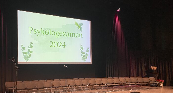 Scen med tomma stolar och en stor skärm med texten "Psykologexamen 2024" och gröna bladdekorationer.