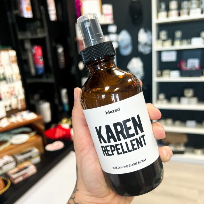 Hand håller en flaska med etiketten "KAREN REPELLENT" framför en hylla med butiksprodukter.