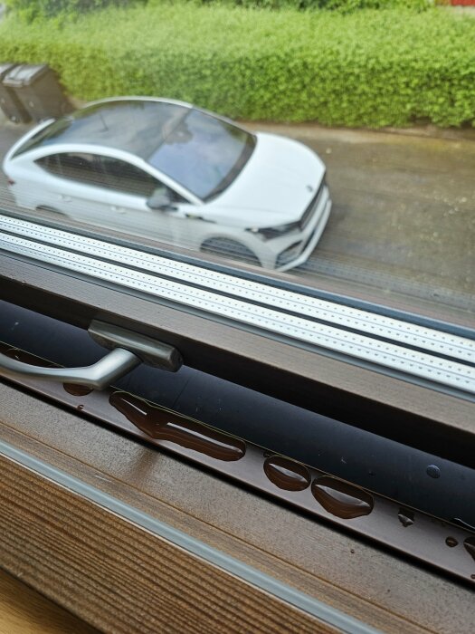 Vattenpölar på inåtslutande brun metallkant vid nedre fönsterkarm med utsikt till parkerad bil.