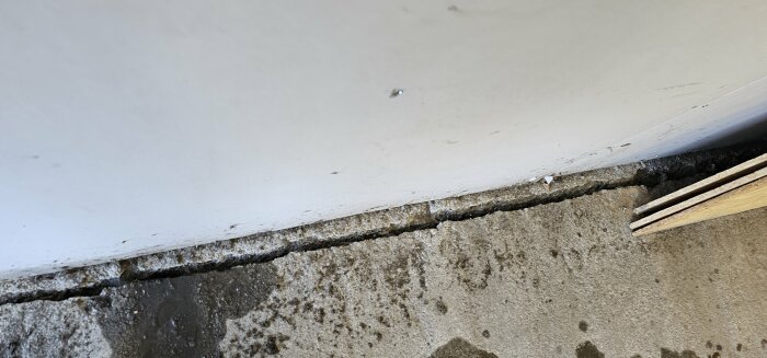 Spricka vid basen av en vitmålad vägg och betongplatta, med synlig skada och lutning, visar behov av reparation.