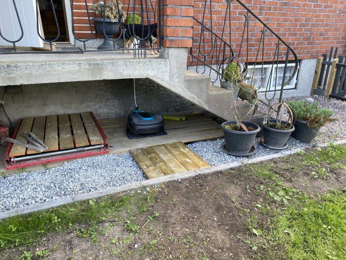 Robotgräsklippare vid laddstation under trapp, bredvid kantsten och blomkrukor på gräsmatta.