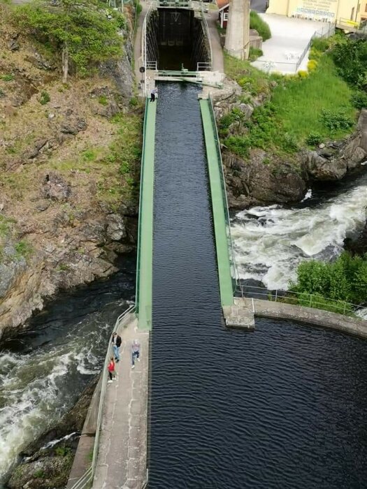 Vy ovanifrån av en grön vattenväg med gångbro längs sidan och människor som promenerar intill forsande vatten.