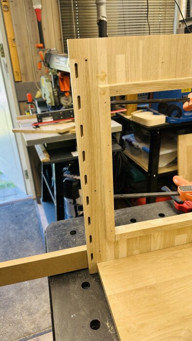 Delar av en träkonstruktion med utfrästa spår för Dominobrickor i verkstadsomgivning.