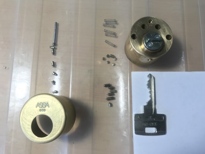 Demonterat ASSA cylinderlås med tillhörande komponenter och nyckel på bord.