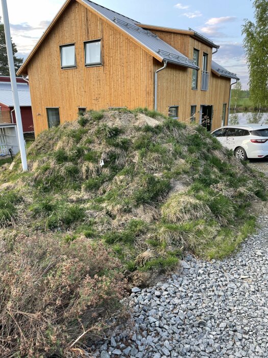 Trädgård på hösten med övervuxen gräskulle framför modernt trähus och parkerad bil.