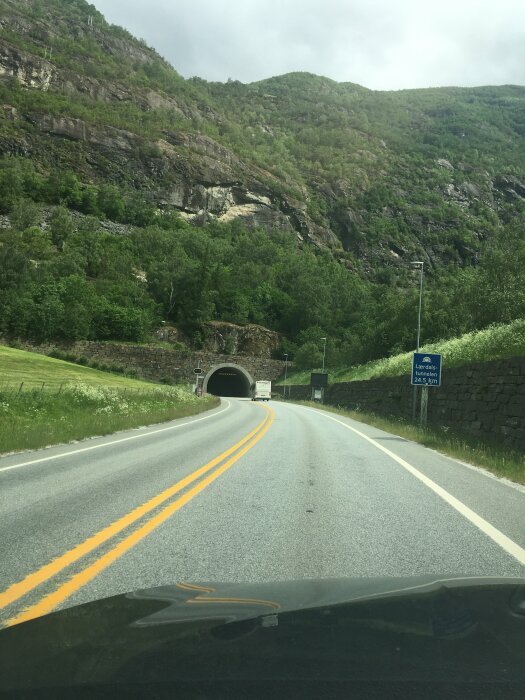 Vy från bil på vindlande väg leder mot en tunnelingång omgiven av gröna kullar och berg.