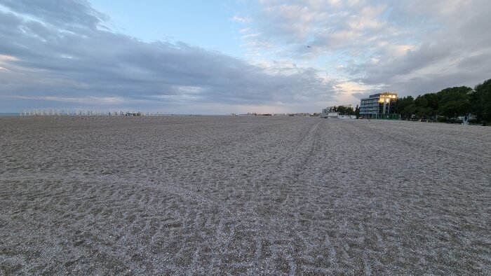 Vidsträckt sandstrand vid skymning med molnig himmel och byggnader i bakgrunden.