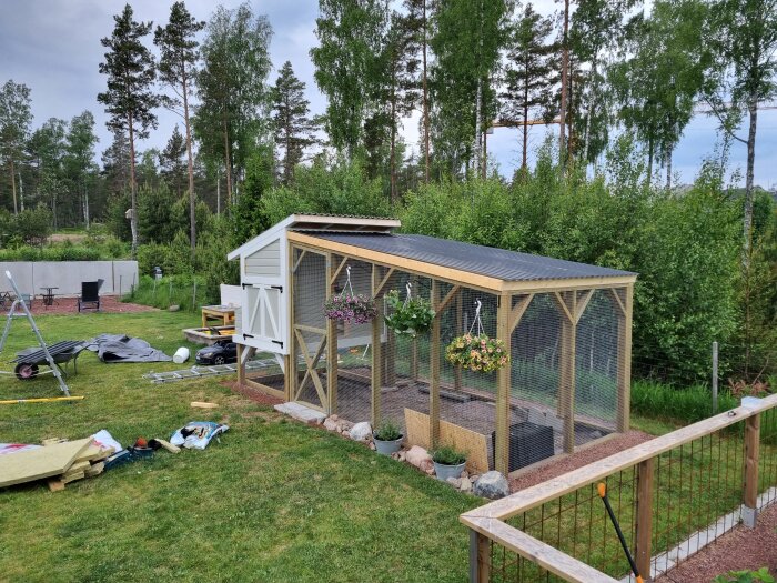 Nybyggd hönsagård i trädgård med vit kycklingkoja, nätad löpgård och blomkrukor, byggmaterial på gräset.