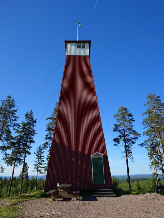 Röd träbrandtorn med svensk flagga på toppen i skogsmiljö mot blå himmel.