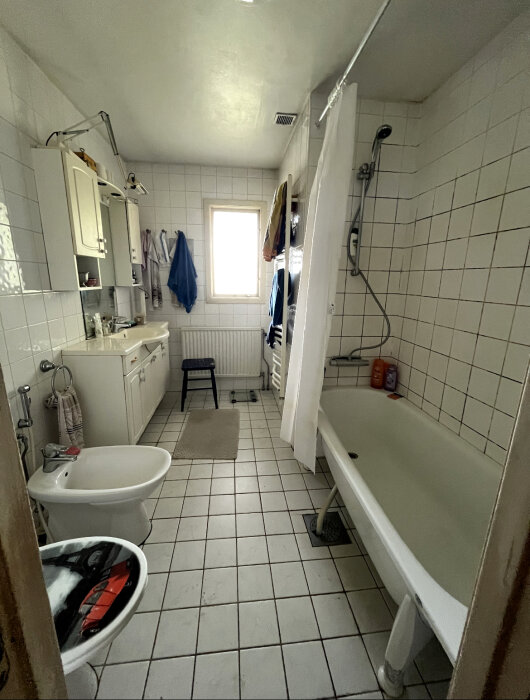 Äldre badrum i behov av renovering, vit kaklad vägg och golv, med badkar, tvättställ, toalett, och handdukar.