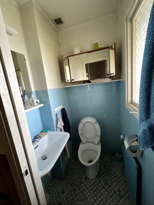 Gammalt badrum med blå kakel, vit handfat, toalett och spegelskåp som behöver renoveras.