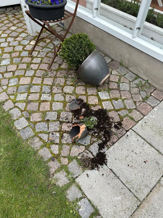 Söndrad blomkruka och jord på stenplattor vid husrad efter en olycka med batteridriven gräsklippare.