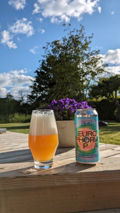 En ölglas fylld med grumlig IPA bredvid en burk märkt "EUROPHORIA IPA" på ett träbord utomhus.