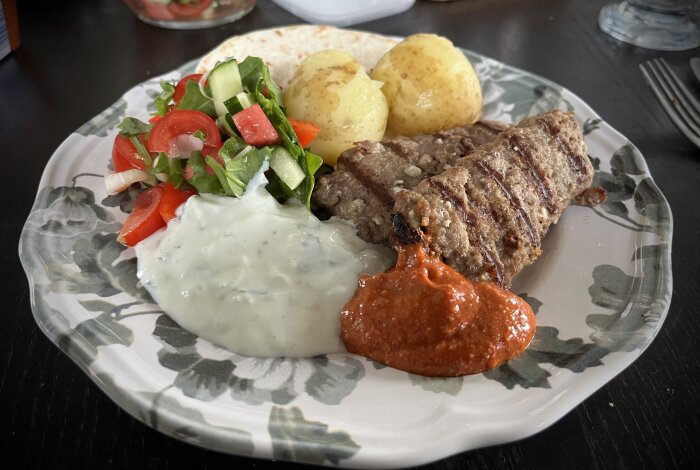 Tallrik med lammfärsspett, färskpotatis, sallad, tsatsiki, tomatröra och minitortilla.