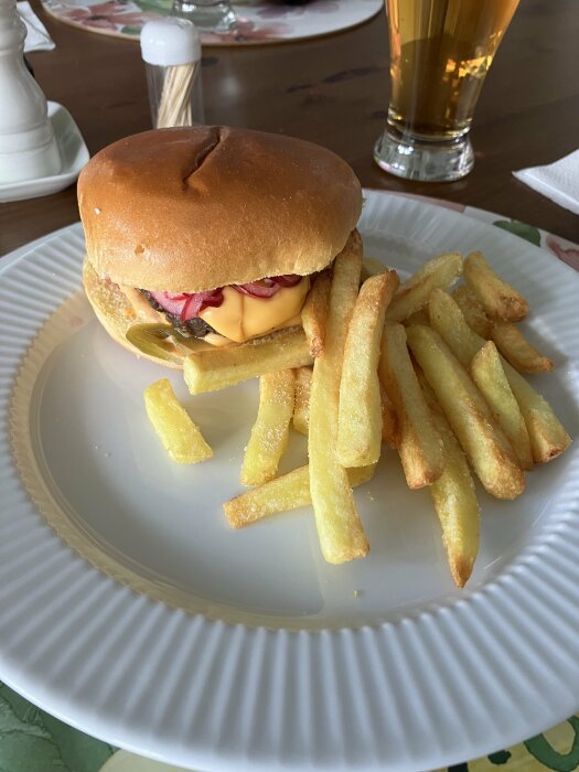 Dubbelburgare med bröd, ost, jalapeños och picklad rödlök serverad med pommes på en vit tallrik, bakgrund med ölglas.