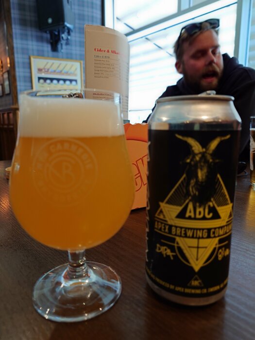 Ölglas med dimmig dryck och burk från Apex Brewing i förgrunden, otydlig person och meny i bakgrunden.