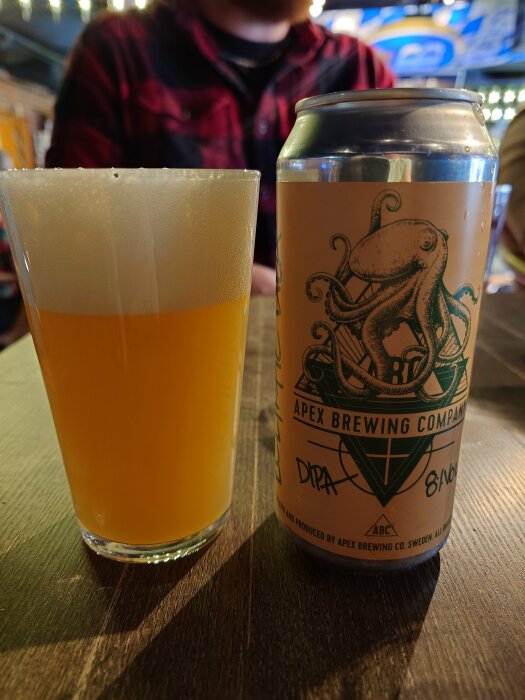 Ölglas fyllt med ljus öl bredvid en Apex Brewing ölburk med en bläckfiskmotiv på ett träbord.