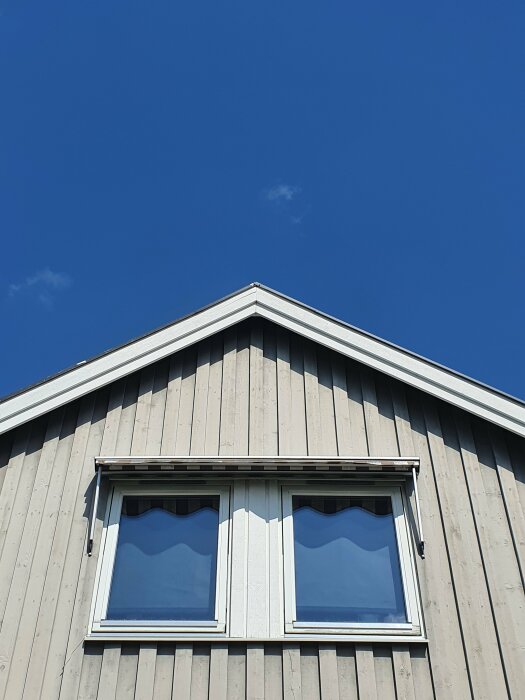 Husfasad med grå panel och vindskivor under en klarblå himmel, framförberedelse för målning.